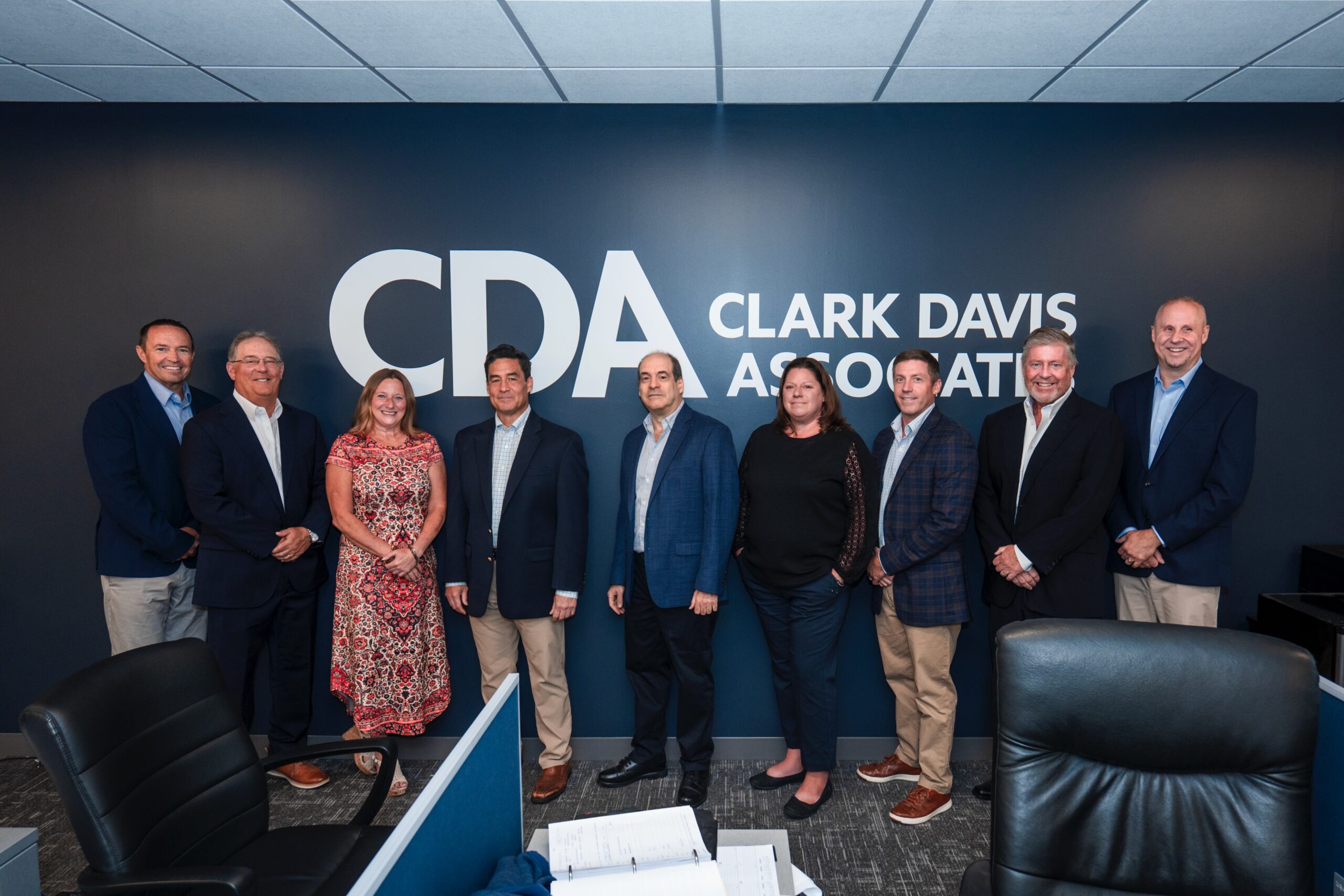 Nine professionals stand together in an office lobby in front of a dark wall featuring the CDA (Clark Davis Association) logo and text, posing for a group photo.